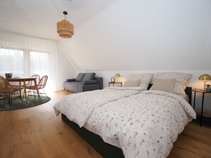 Et soveværelse med en stor seng, en grå sofa, et spisebord med stole og en hængende lampe.
