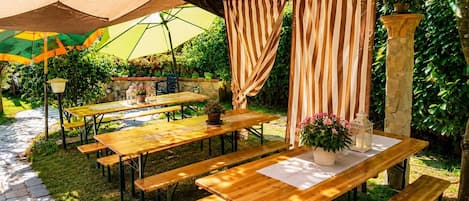 An outdoor patio area with wooden tables and benches, covered by a wooden pergola, and surrounded by lush greenery.