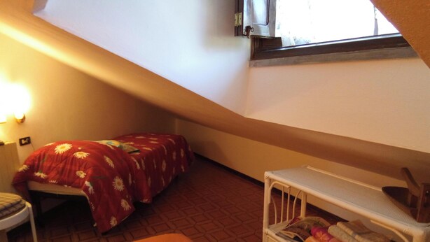 Une petite pièce agréable, avec un plafond en pente, un lit simple recouvert d’une courtepointe à motifs floraux, une petite table et une cage pour animaux de compagnie.