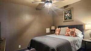 A bedroom with a bed, a ceiling fan, a wall-mounted picture, and a nightstand with a lamp.