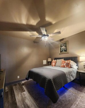 A bedroom with a bed, a ceiling fan, a wall-mounted picture, and a nightstand with a lamp.