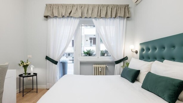 A bedroom with a large bed, a window with curtains, a radiator, and a small table with a lamp.