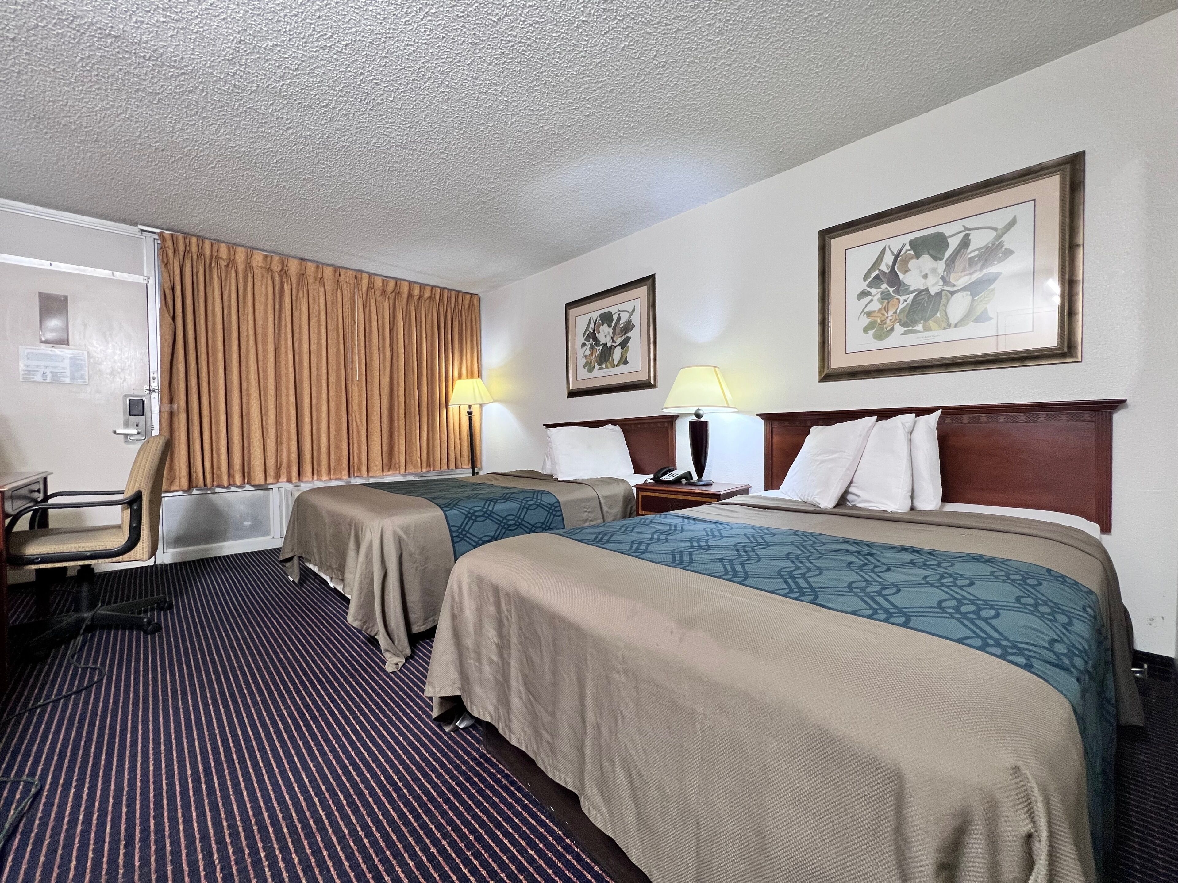 A hotel room with two beds, a desk, a chair, a lamp, and a picture on the wall.