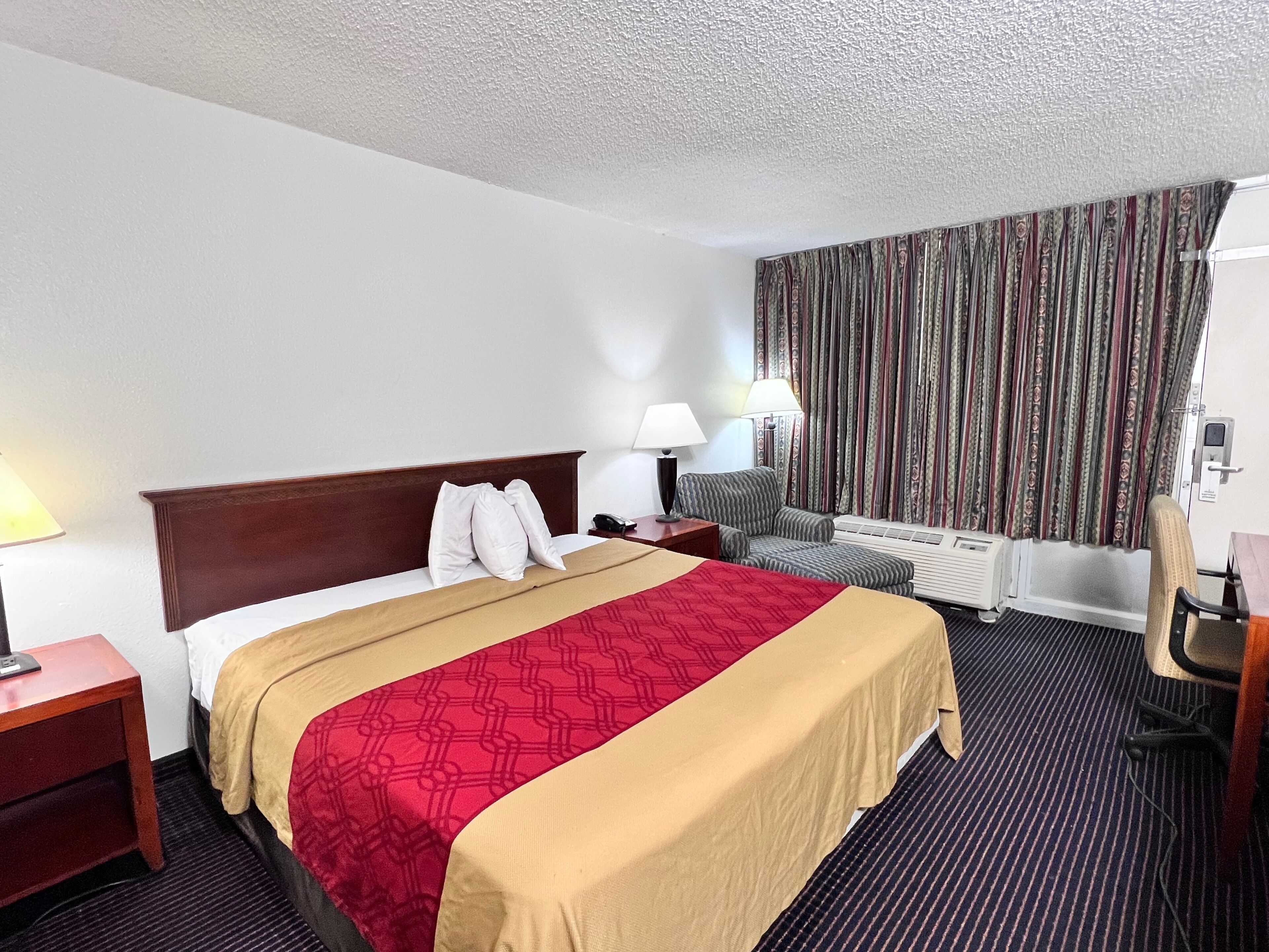 Habitación de hotel con una cama grande, un escritorio, una silla, una lámpara y una ventana con cortinas.