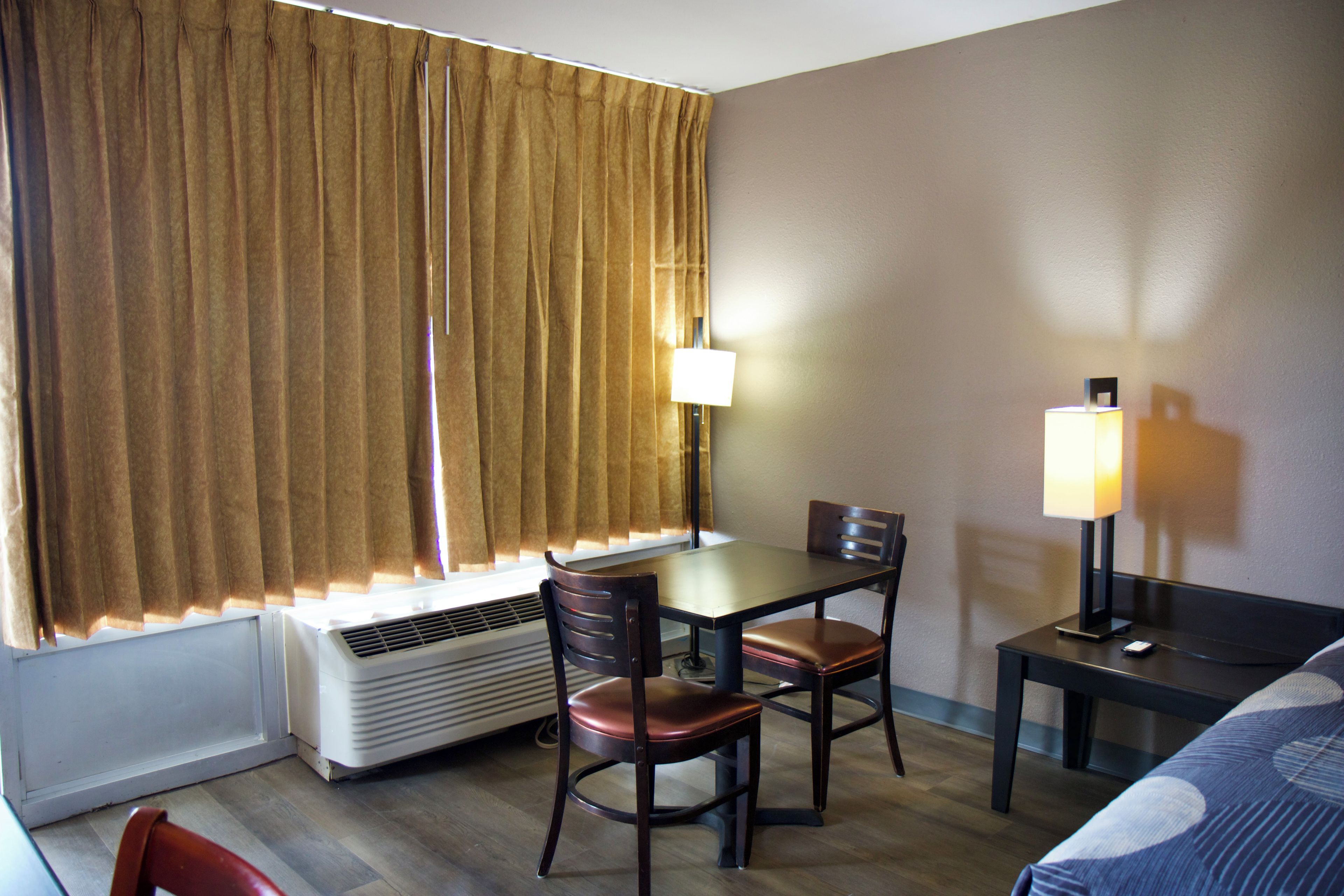 A hotel room with a bed, a desk with a chair, a lamp, a heater, and a window with curtains.