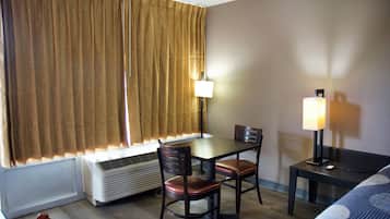 A hotel room with a bed, a desk with a chair, a lamp, a heater, and a window with curtains.