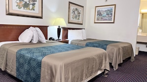 A hotel room with two beds, a nightstand with a lamp, and framed botanical artwork on the walls. - Heritage Inn and Suites (Baton Rouge)