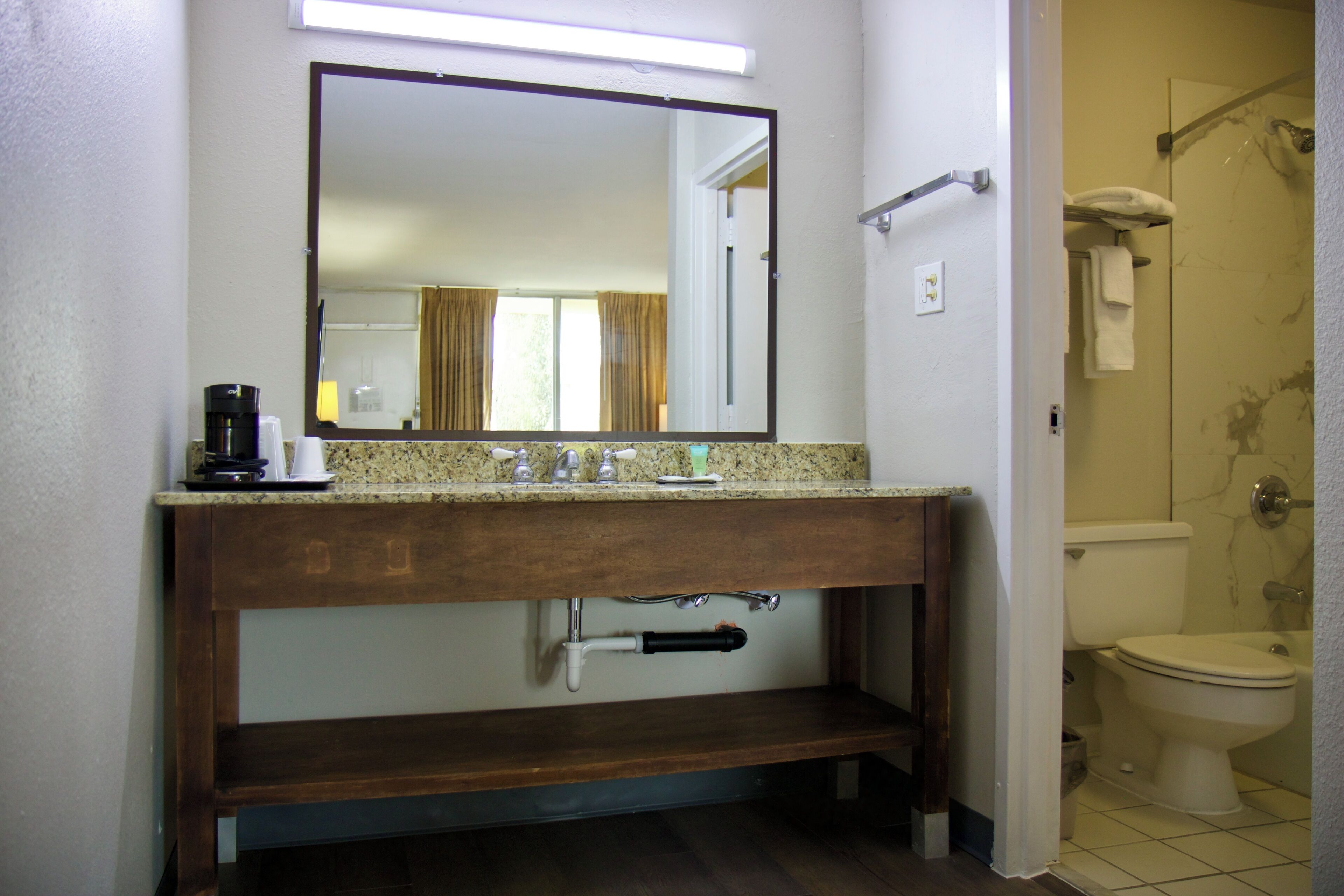 A bathroom with a wooden vanity, a mirror, a coffee maker, and a toilet visible through an open door.
