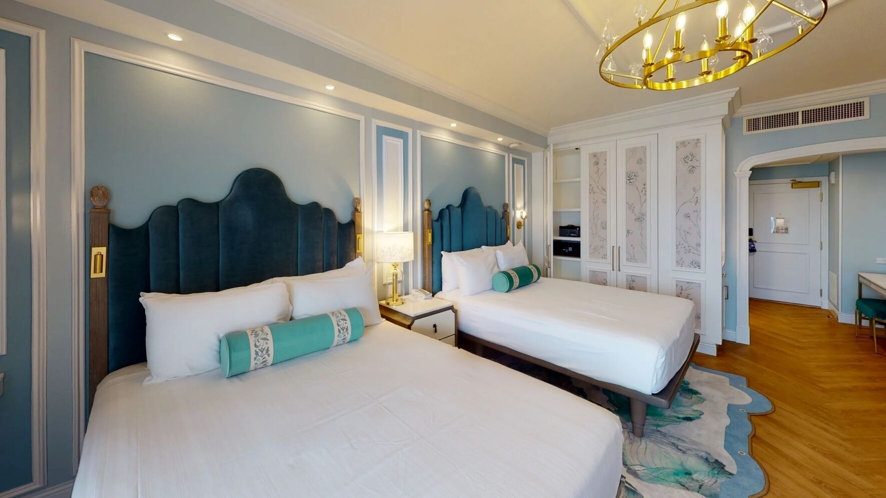 A hotel room with two beds, a chandelier, a desk, and a wardrobe.