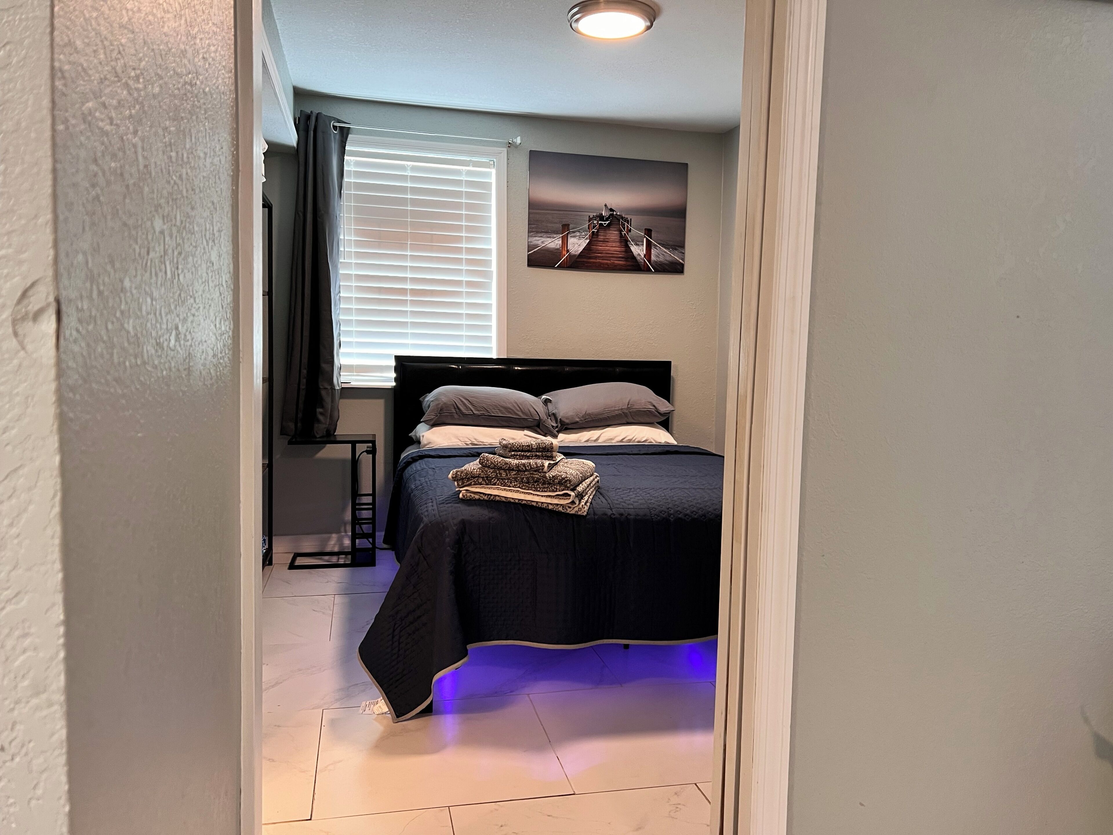 Un dormitorio con cama, ventana con persianas, un cuadro en la pared y una lámpara de techo.