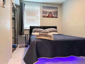 A bedroom with a bed, a window with blinds, a painting on the wall, and a blue rug. - Cozy 1-bedroom apartment in perfect Clearwater with AC (Clearwater)