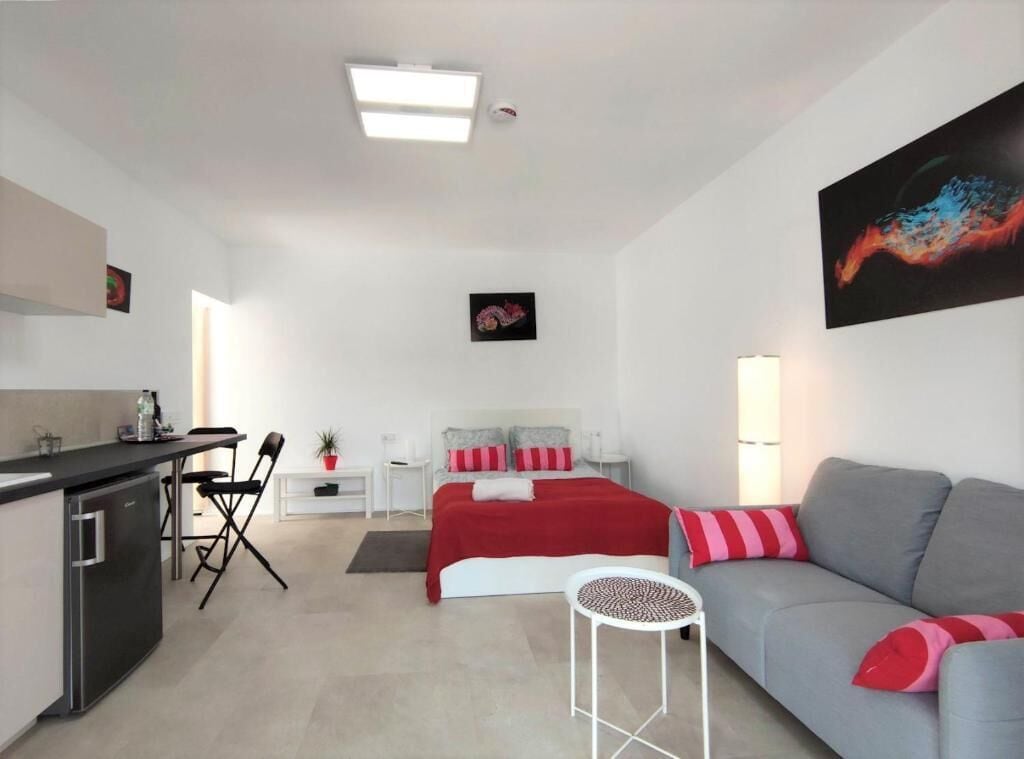 A modern hotel room with a kitchenette, a bed with red bedding, a grey sofa, and a painting on the wall.