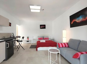 Ein modernes Hotelzimmer mit Küchenzeile, einem Bett mit roter Bettwäsche, einem grauen Sofa und einem Bild an der Wand.