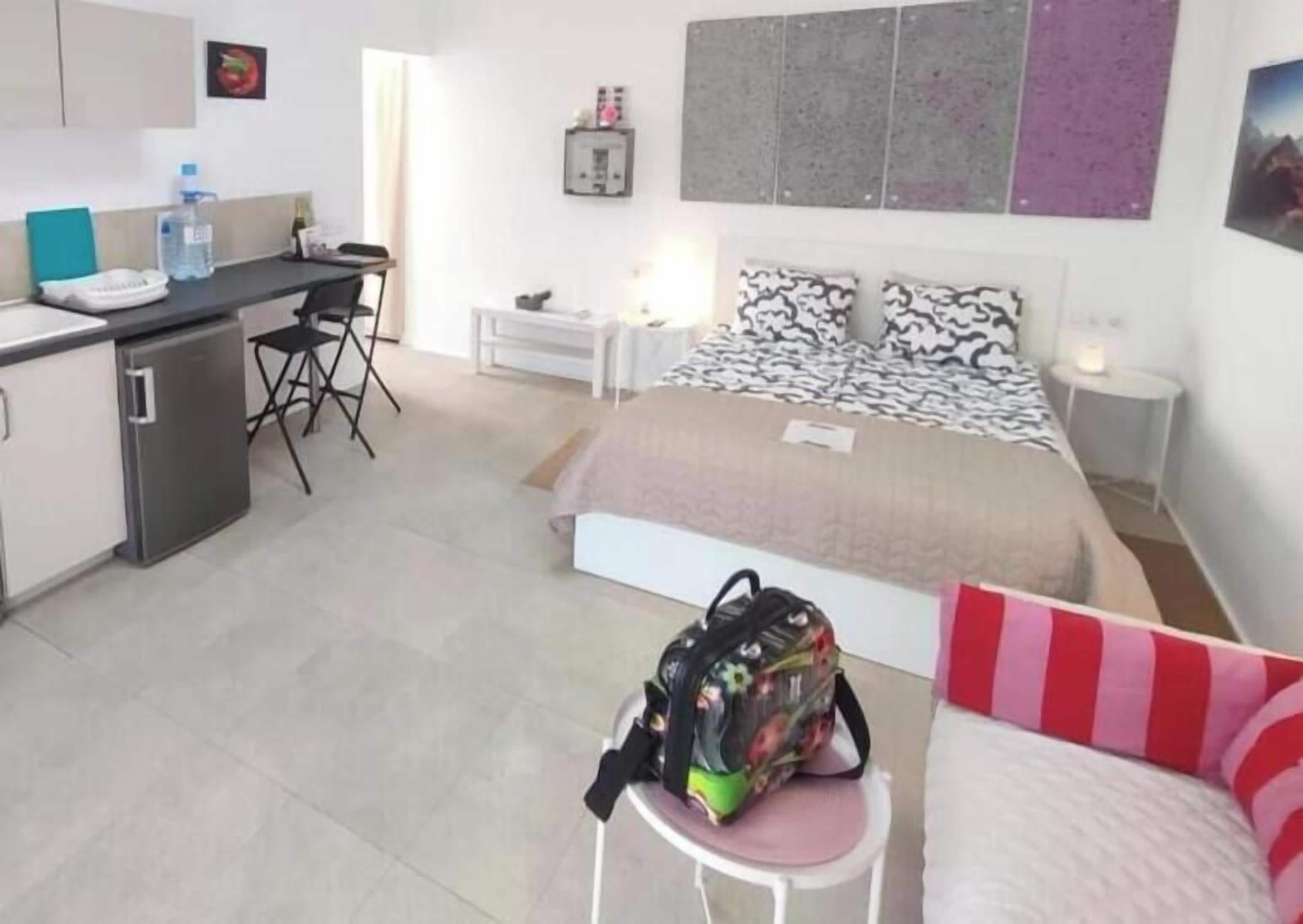A compact hotel room with a kitchenette, a bed with patterned bedding, a small seating area, and a desk.