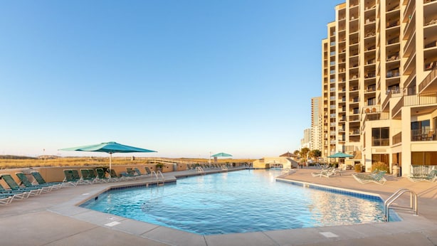 Piscina com espreguiçadeiras e guarda-sóis, cercada por um edifício alto.