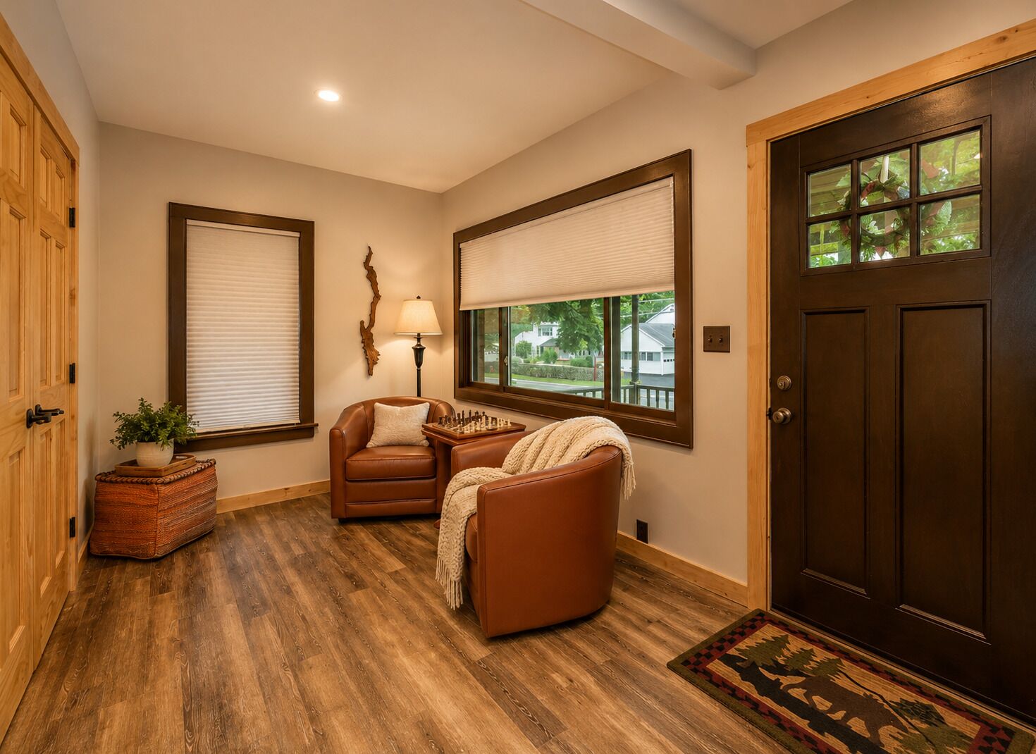 Una habitación con piso de madera, un sillón de cuero marrón, un armario de madera con una planta, una lámpara y una puerta con ventanal.