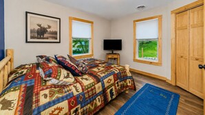 Ein Schlafzimmer mit einem großen Bett, zwei Fenstern mit Jalousien, einem Fernseher auf einem Holzboden und einer Tür.