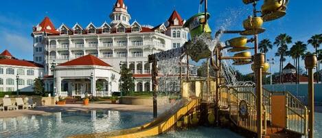 A hotel with a large pool, a water fountain with golden cups, and a grand building with multiple balconies and a red roof.