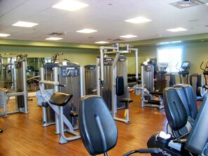 A gym with various exercise machines, including a leg press and a chest press, with a wooden floor and a green wall in the background. - Orlando | Disney Magic Experience | Pool + Spa (Lake Buena Vista)