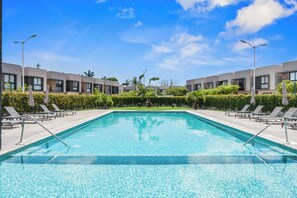 A rectangular swimming pool with a tiled edge, surrounded by lounge chairs and umbrellas, with modern buildings in the background. - Casa Ivarica Spacious Home for 16 in Residential (Mezcales)