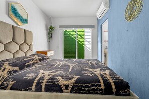 A bedroom with a large bed, a wall-mounted air conditioner, a window with a view of a green lawn, and a decorative wall piece. - Casa Ivarica Spacious Home for 16 in Residential (Mezcales)