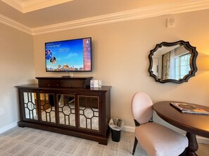 A hotel room with a television displaying Disney Springs, a dark wooden cabinet with glass doors, a round table with a magazine, and a mirror on the wall. - Orlando | Elegant Short Stay | Pool + Spa (Lake Buena Vista)