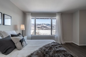 A bedroom with a bed, a lamp, a window with curtains, and a view of mountains. - Le Paradis Tranquille: Beautiful Mountain Views! (Mont-Tremblant)