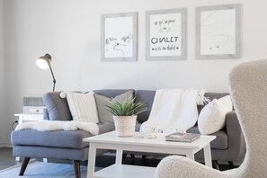 Ein modernes Wohnzimmer mit einem grauen Sofa, einem weißen Couchtisch und einer Stehlampe. An der Wand hängen drei gerahmte Kunstwerke.