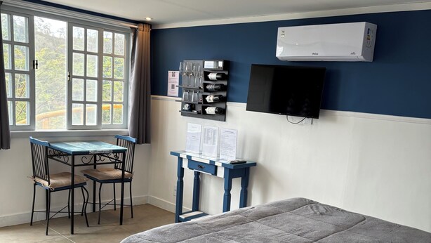 A bedroom with a bed, a desk, and a chair. There is a window with curtains, a flat-screen TV mounted on the wall, and a wall-mounted air conditioner.