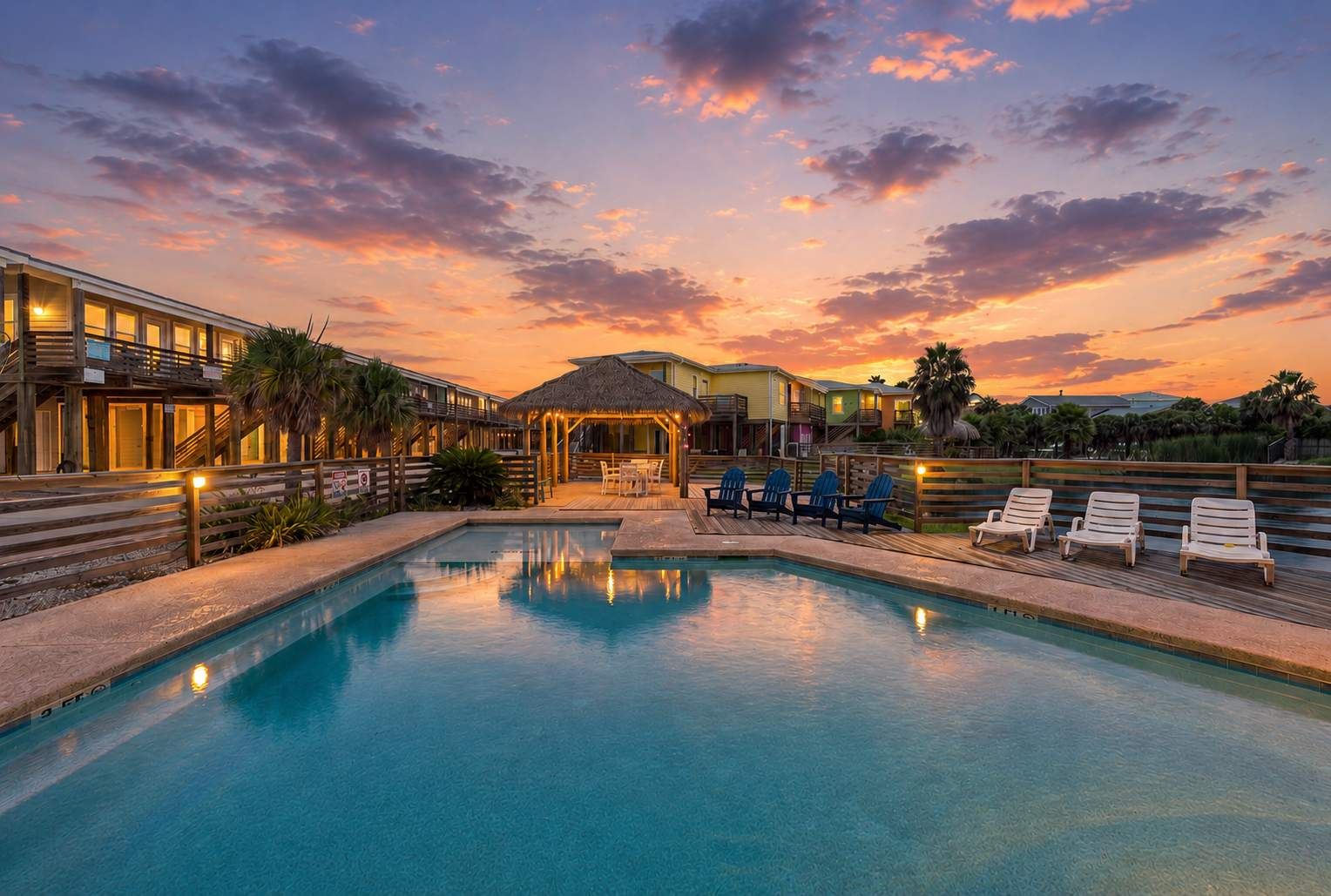 A swimming pool with lounge chairs and a thatched-roof structure, surrounded by a wooden deck and palm trees.