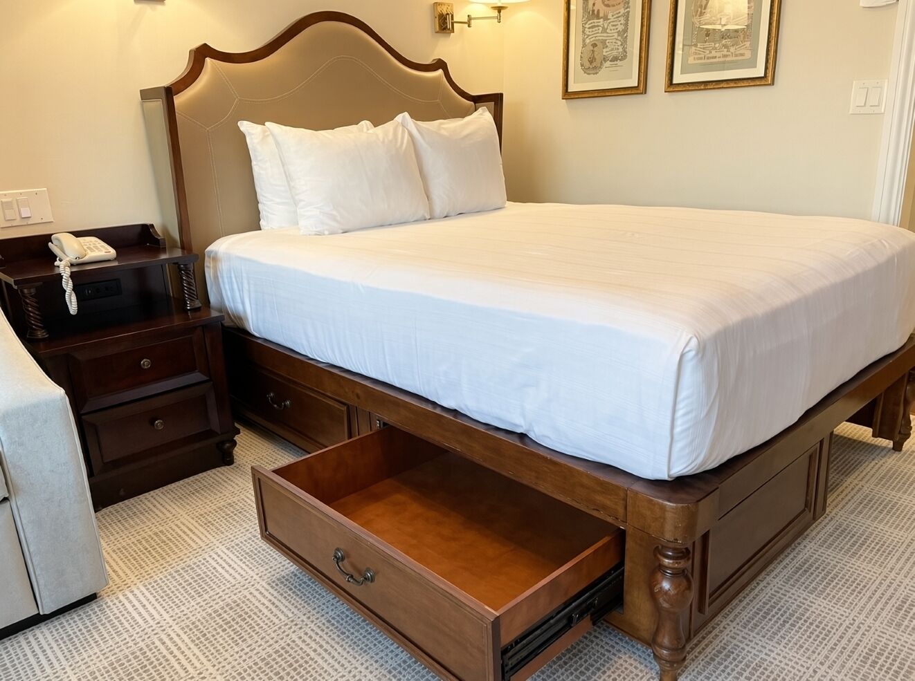 A hotel room with a large bed, a bedside table with a phone, a drawer, and two framed pictures on the wall.