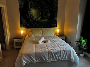 A neatly made bed with a rolled towel, two bedside lamps, a large forest-themed tapestry on the wall, and a potted plant to the side. - Moose - Private Bedroom near Downtown and 17th Av (Calgary)