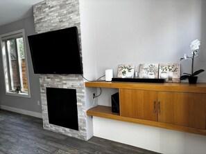 Ein modernes Wohnzimmer mit einem flachbildfernseher über einem Kamin, Holzkästen darunter und Dekorationen auf dem Kaminvorsatz.