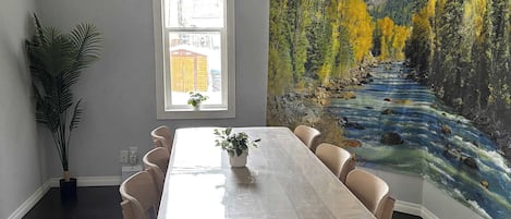 Ruokailutila, jossa on pöytä ja tuolit, ikkuna, kasvi ja seinämaalaus, jossa on kuvattuna joki ja vuoristo.