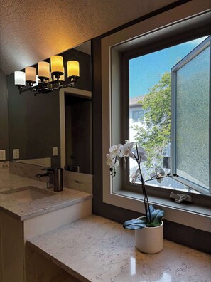 Une salle de bain dotée d’un plan de toilette en marbre, d’un lavabo, d’un miroir, d’une fenêtre donnant sur un espace verdoyant et d’un luminaire fixé au mur.