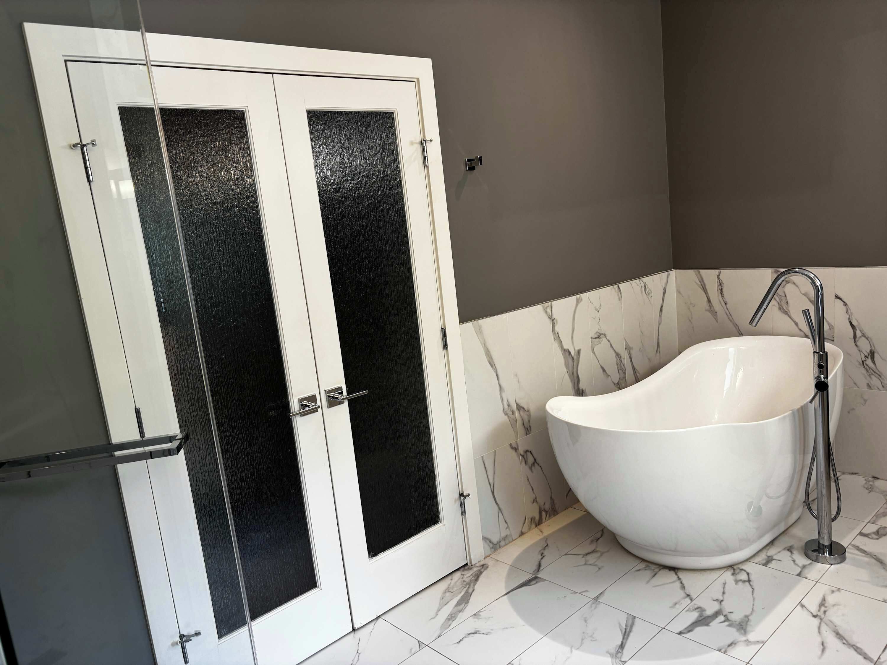 A bathroom with a white freestanding bathtub, a double door with a frosted glass panel, and marble-patterned tiles.