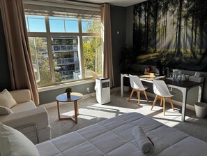 Une chambre d’hôtel comprenant un lit, un bureau, deux chaises, une petite table et une grande fenêtre donnant sur des immeubles et des arbres.