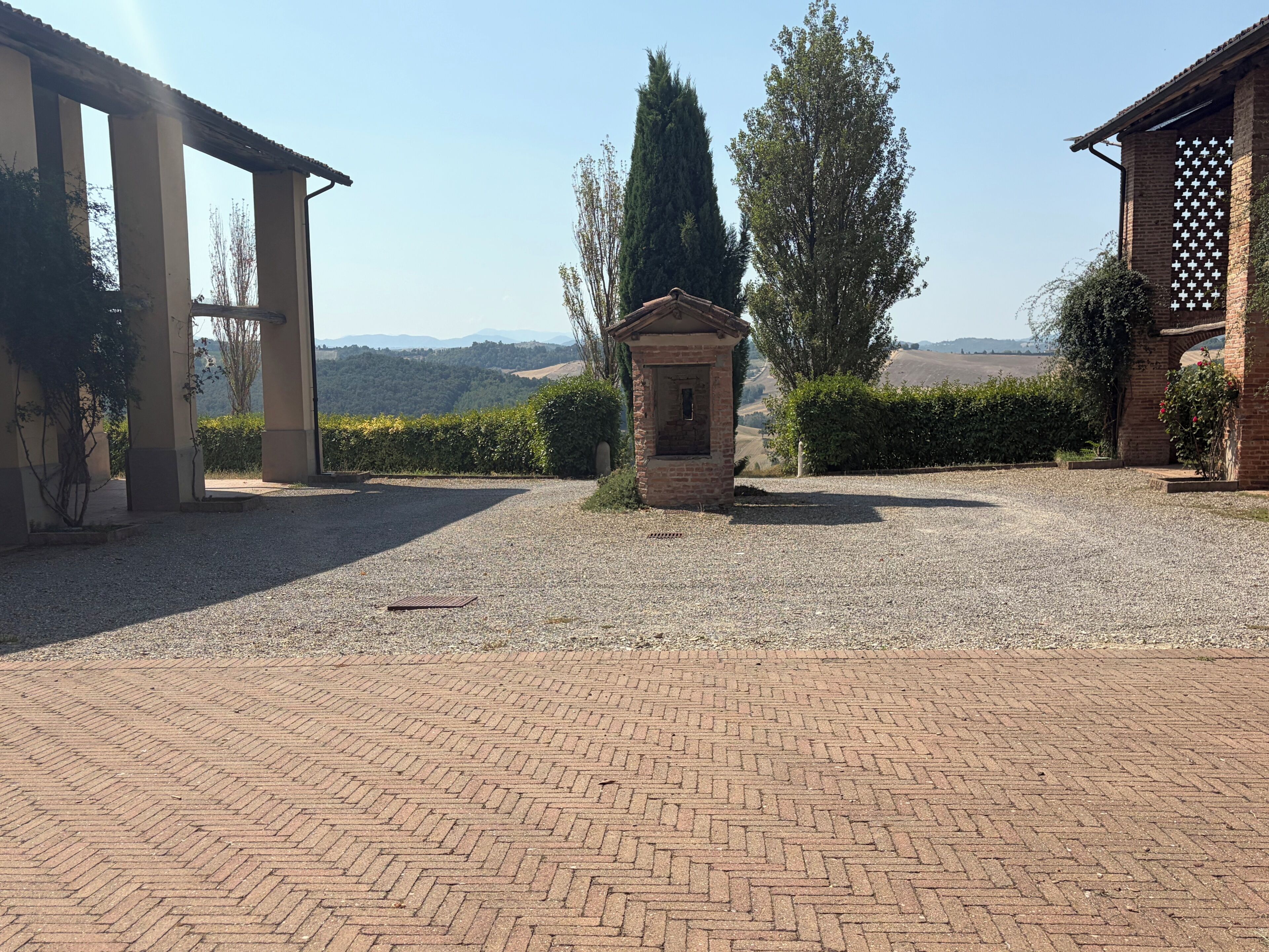A gravel courtyard with a brick pathway leading to a small brick structure, surrounded by greenery and a view of distant hills.