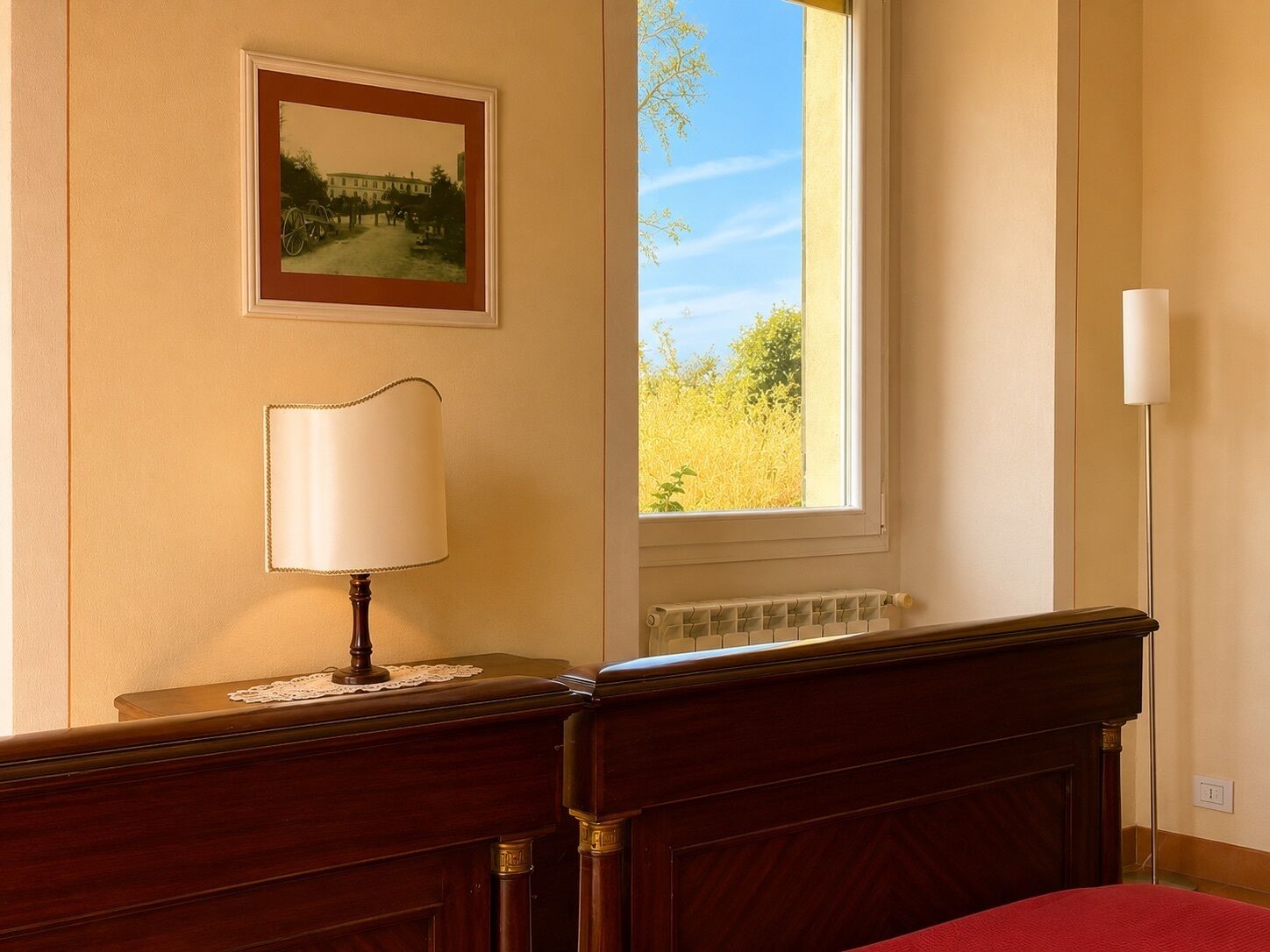 Una habitación con una cama, una mesita de noche con una lámpara, un cuadro en la pared y una ventana con vistas a la vegetación.