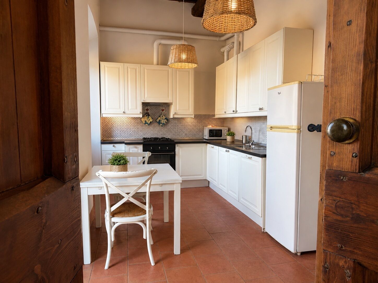 Une cuisine avec des meubles blancs, un plan de travail noir, une table blanche avec une chaise, un four à micro-ondes et un réfrigérateur.