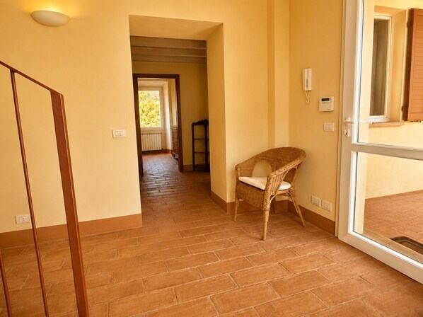 A hallway with a wicker chair, a wooden door frame, and a window with a view of greenery.