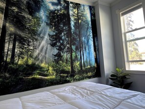 Ein Schlafzimmer mit einer großen Wandteppich-Szene aus Waldlandschaft, einem Bett mit weißer Bettwäsche und einem Fenster mit natürlichem Licht.