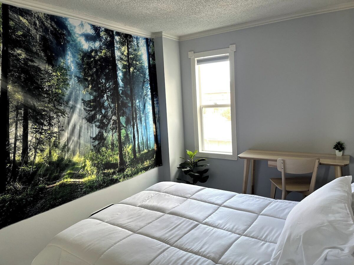 A bedroom with a bed, a desk, a chair, a window, and a forest mural on the wall.