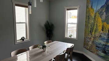 Une salle à manger avec une longue table, quatre chaises, deux fenêtres équipées de stores, une plante en pot et un grand tableau représentant un paysage accroché au mur.