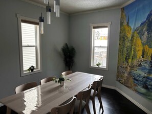 A dining room with a long table, four chairs, two windows with blinds, a potted plant, and a large landscape painting on the wall. - Bear - Private Master Bedroom near DT and 17th Av (Calgary)