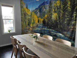 A dining area with a table and chairs, a window with a view of a building, and a mural of a river and forest. - Wolf - Private Bedroom near Downtown and 17th Av (Calgary)