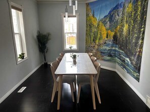 Un espace repas comprenant une longue table, des chaises, une plante en pot et une grande fresque représentant une forêt et une rivière.