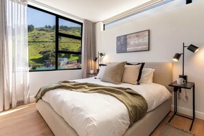Ein Schlafzimmer mit einem großen Fenster, einem Bett, Nachttischlampen, einem gerahmten Bild und einem Nachttisch.