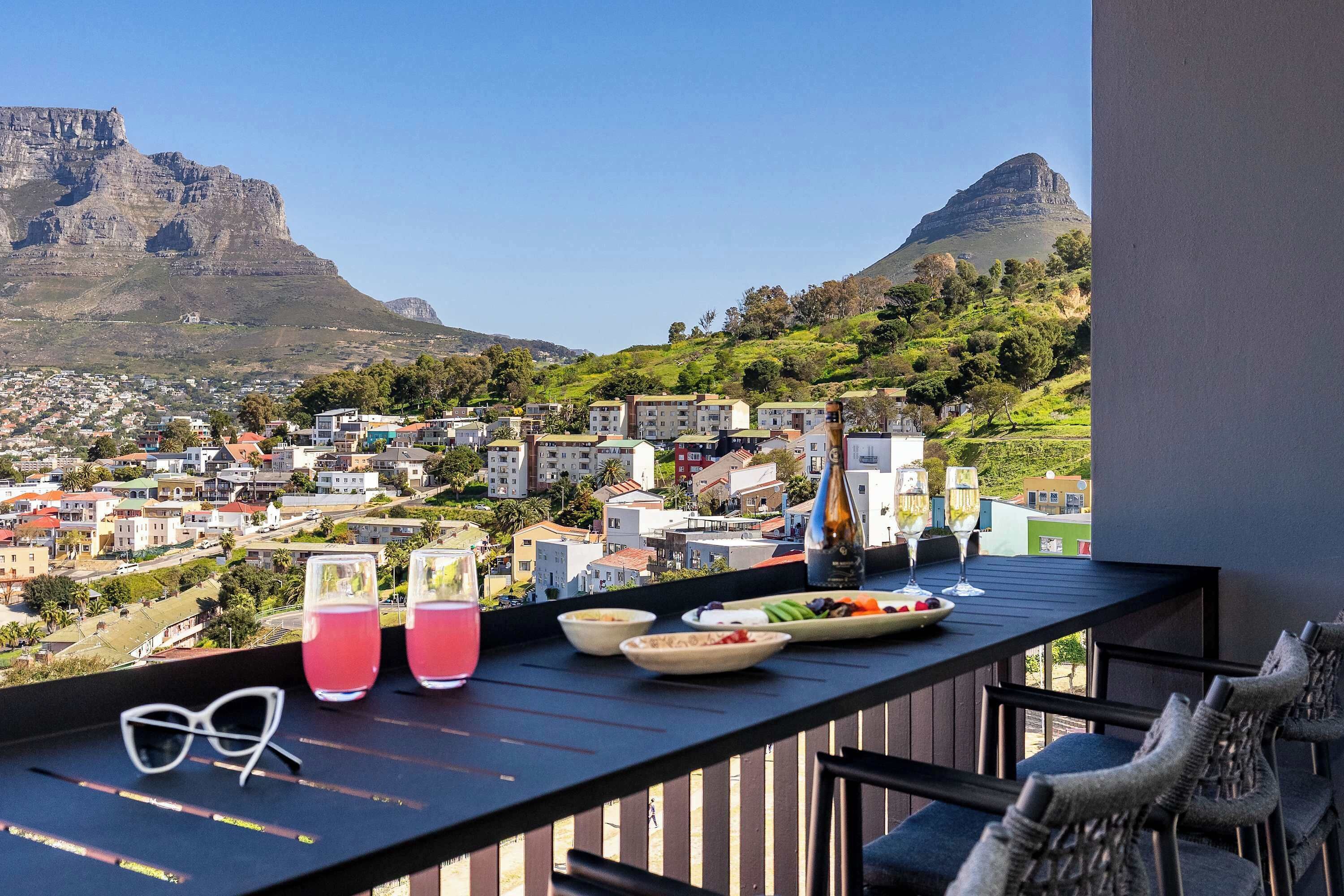 A balcony with a table set for dining, glasses, and a bottle, overlooking a town with buildings and mountains.