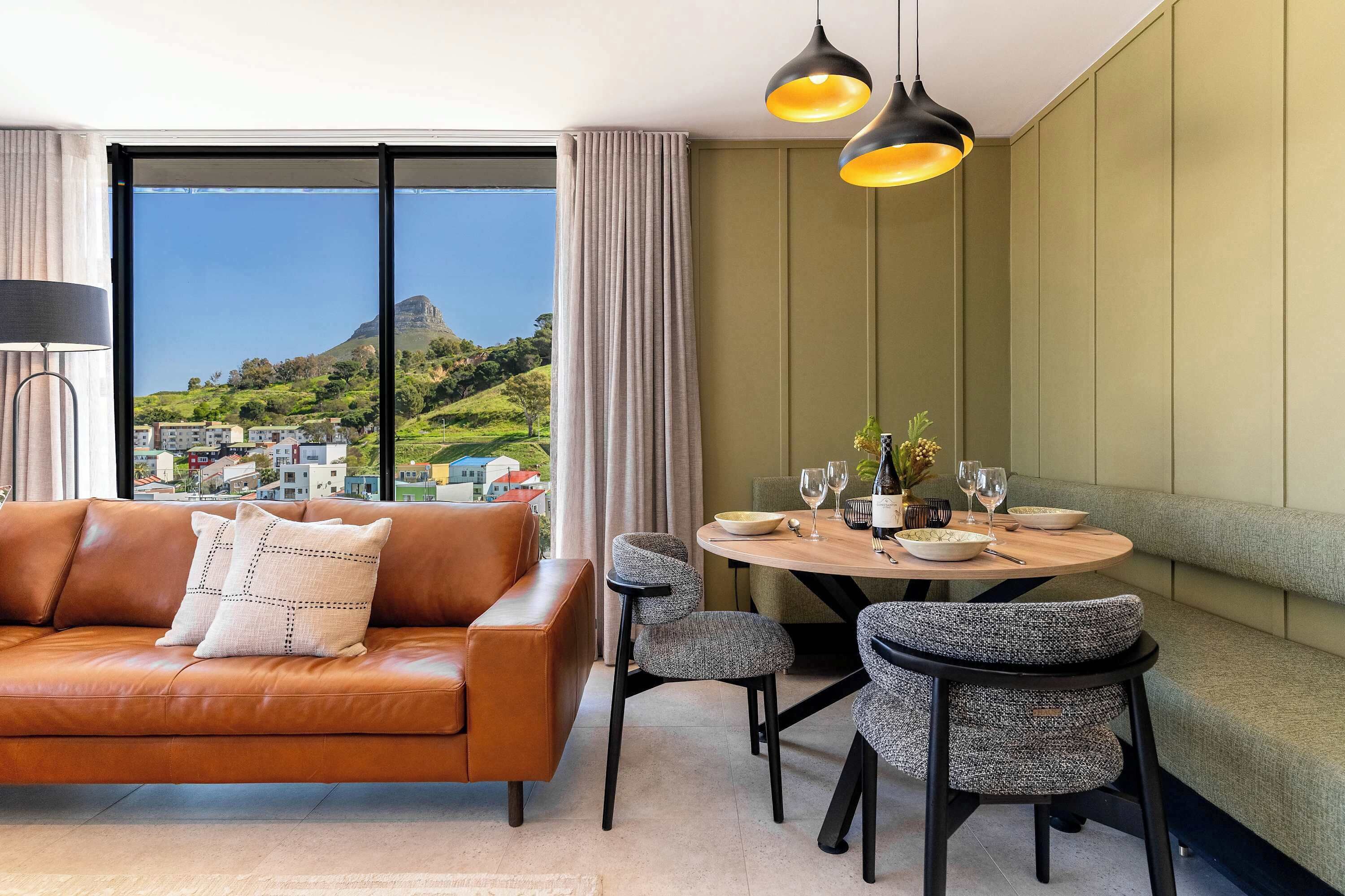 Un salon moderne avec un canapé en cuir, une table à manger et des chaises, et une vue sur un paysage montagneux grâce à de grandes fenêtres.